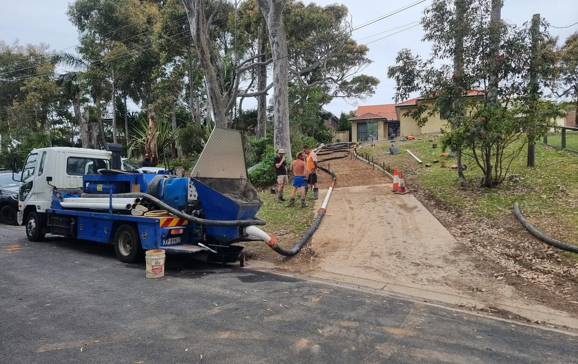 Concrete pump hose running across a grassed rural yard from the truck to a house slab pour, surrounded by tall gum trees