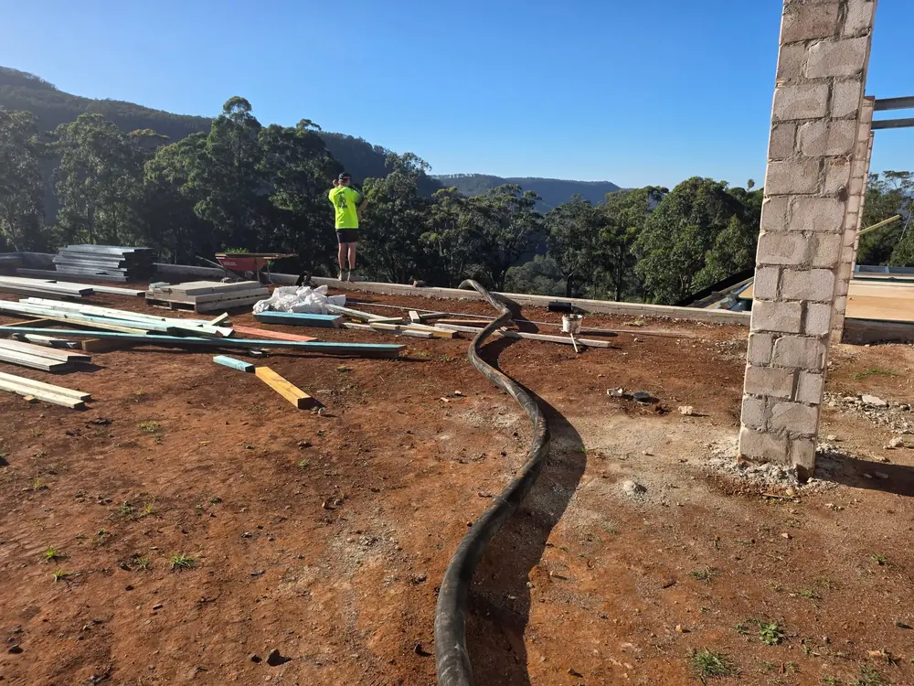 Worker screeding a freshly poured concrete slab beside a retaining wall, with hills in the background